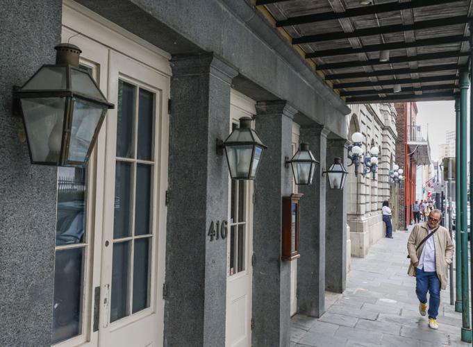 A man walks down a sidewalk next to a historic building with large lanterns and columns. The street is lined with more buildings in the background. Hes wearing a beige jacket, jeans, and carrying a bag.