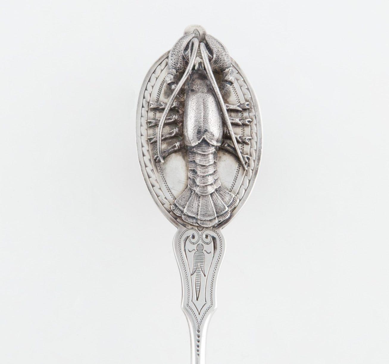 An ornate silver spoon with an intricately detailed design of a lobster on the handles bowl, set against a plain white background.