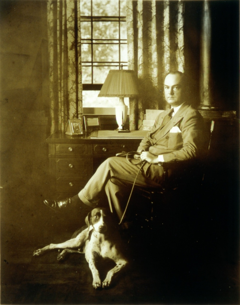 A sepia-toned image of a man in a suit sitting on a chair next to a wooden desk with a lamp and framed photo. He holds a leash attached to a dog lying on the floor beside him. A window with curtains is visible in the background.