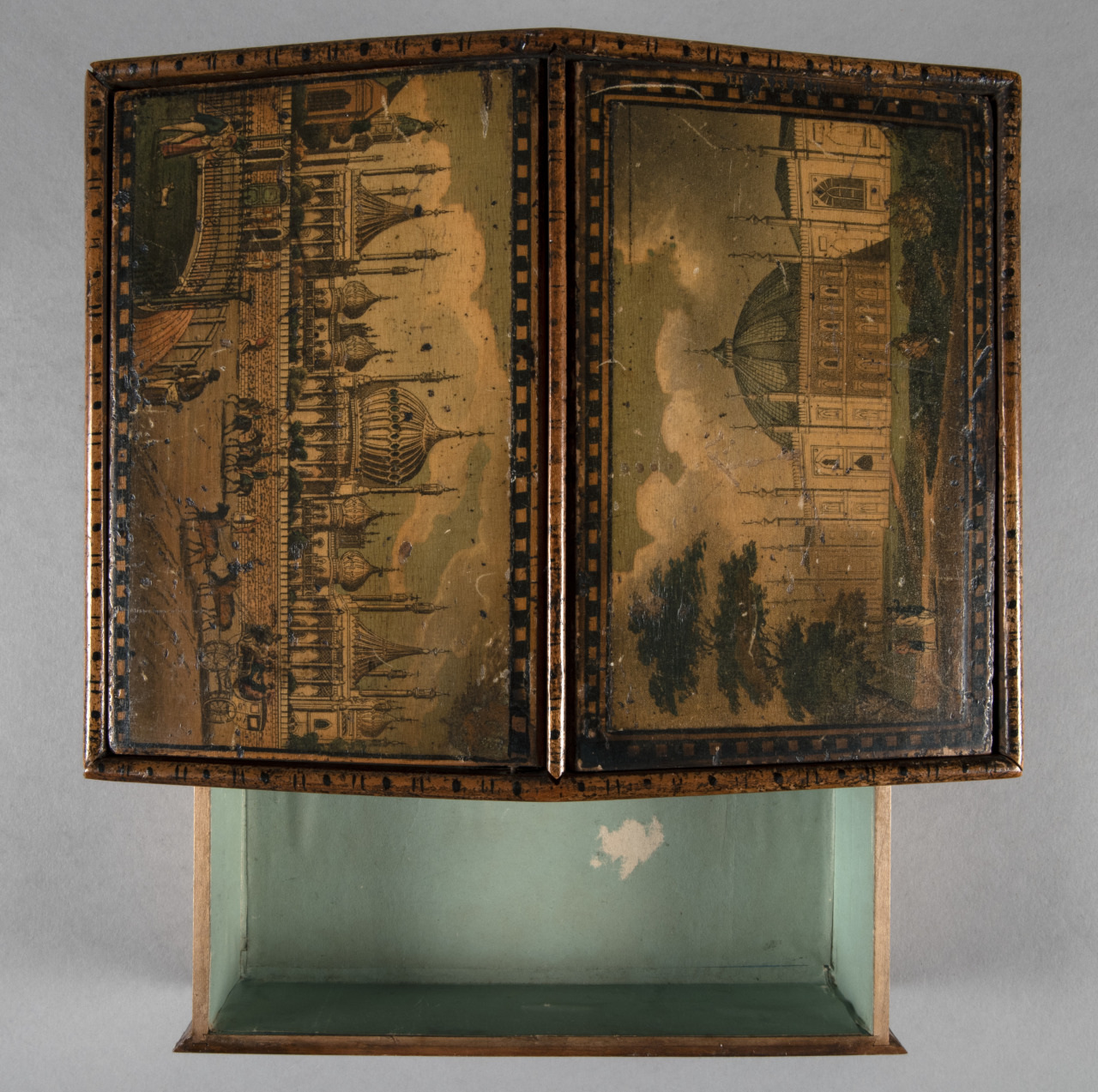 An antique wooden box with hinged doors, displaying intricate scenes of grand architecture. The box is open, revealing a light green interior. The exterior paintings feature detailed buildings and trees, with a vintage, ornate style.
