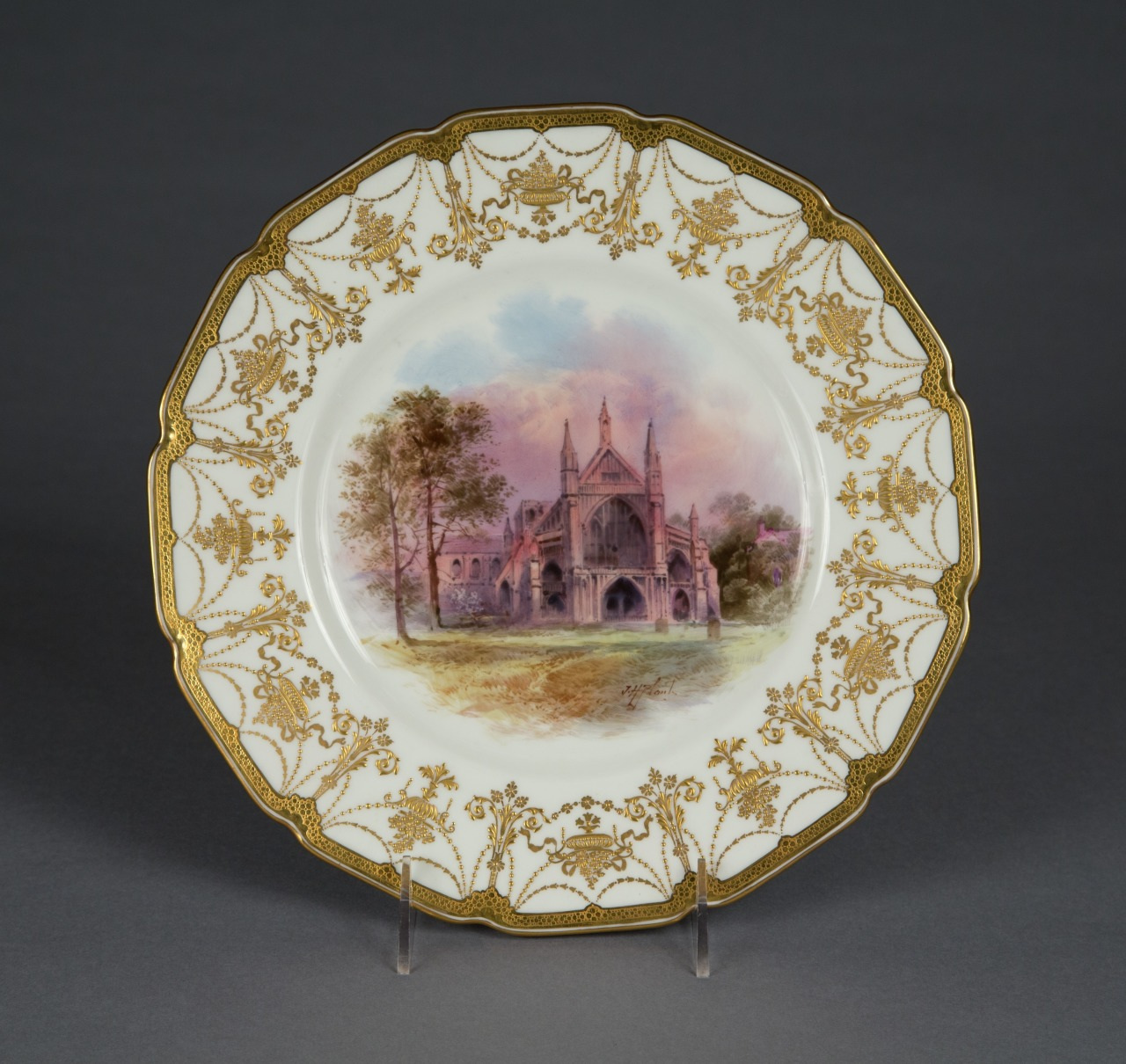 A decorative porcelain plate featuring an intricate gold filigree border and a detailed painting of a Gothic cathedral surrounded by trees under a vibrant sky. The plate is displayed on a stand against a dark background.