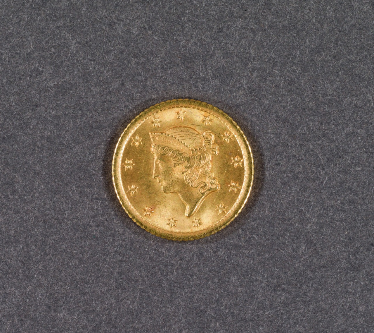 A gold coin featuring a left-facing profile of a woman wearing a headband. Stars surround the profile along the coins edge, set against a textured gray background.