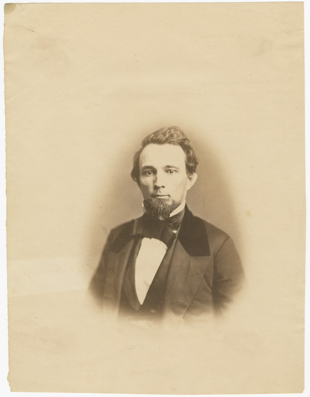 A sepia-toned vintage portrait of a man with styled hair and a beard, wearing a dark suit and bow tie. The image has an aged appearance, with a plain background.