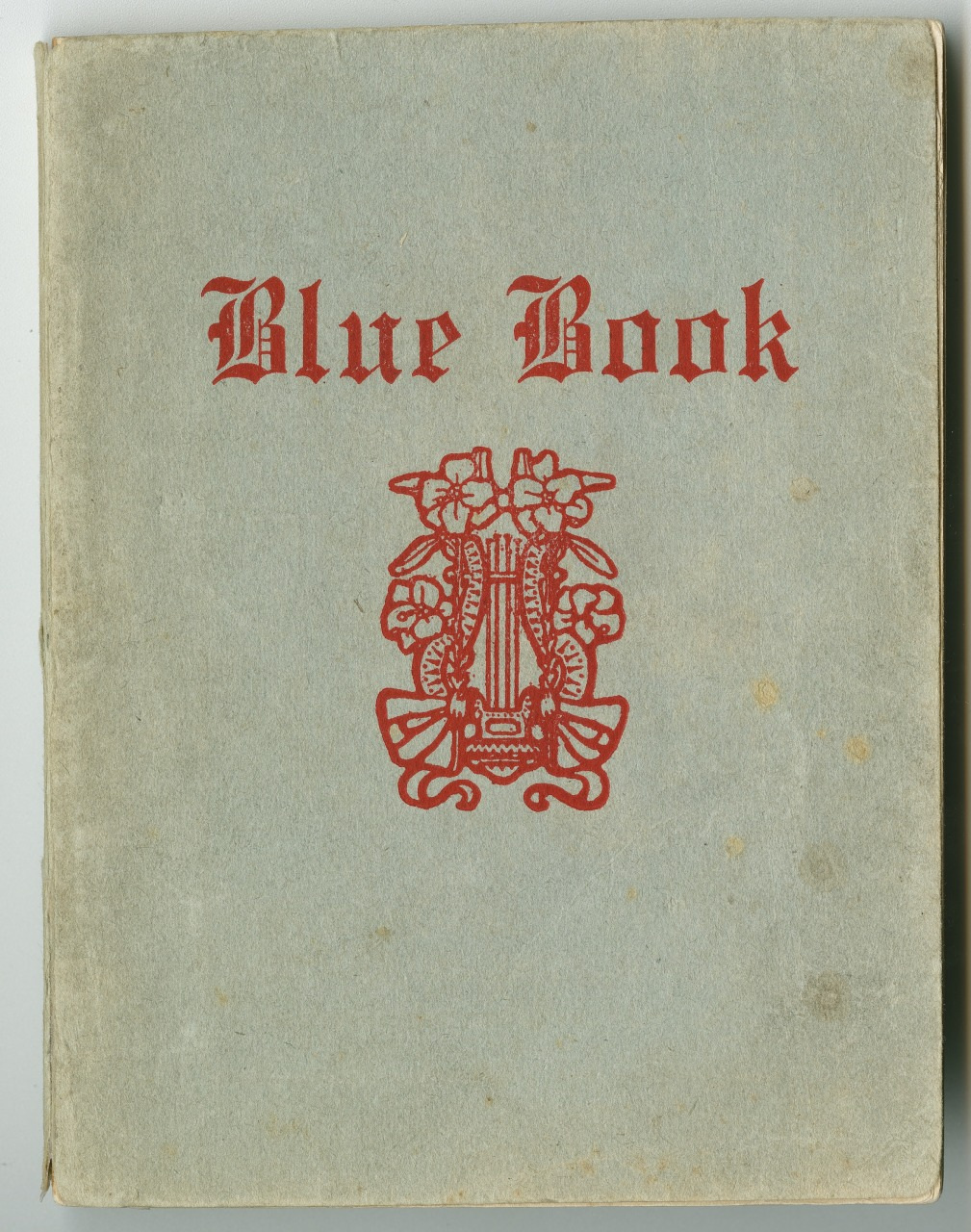 A vintage book cover featuring the title Blue Book in red gothic font. Below the title, there is an ornate red emblem with musical motifs on a textured, pale blue background.