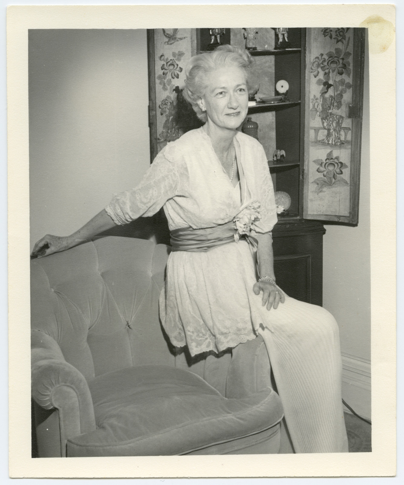An elderly woman with white hair sits beside a plush armchair, wearing a light-colored dress with a floral accessory. A cabinet with decorative items is in the background. She has a calm expression and rests one hand on the chair.