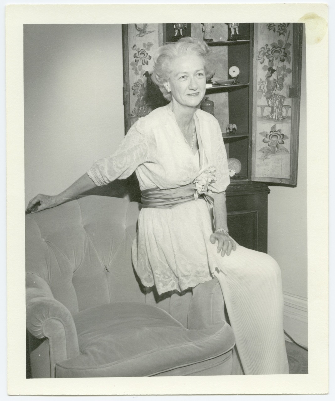 An elderly woman in a light-colored dress stands beside a vintage armchair, with one hand resting on it. Behind her is a cabinet with various decorative items. The setting appears to be a classic, elegantly furnished room.