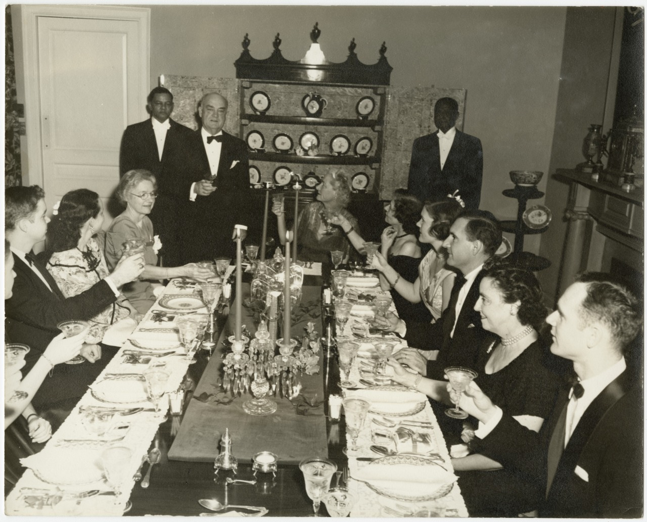 A black and white photo of a formal dinner party. A group of people dressed in formal attire sit around a long, elegantly set table. One man stands, raising a glass for a toast, while others join in, surrounded by vintage decor and china cabinets.