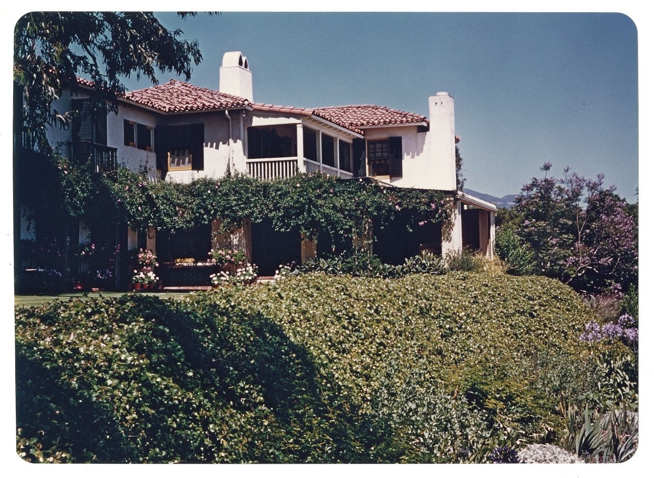 A two-story house with a red-tiled roof and white stucco walls is adorned with lush greenery and flowering plants. A hedge runs along the property, and the house features a spacious balcony with a pergola. A clear blue sky is in the background.