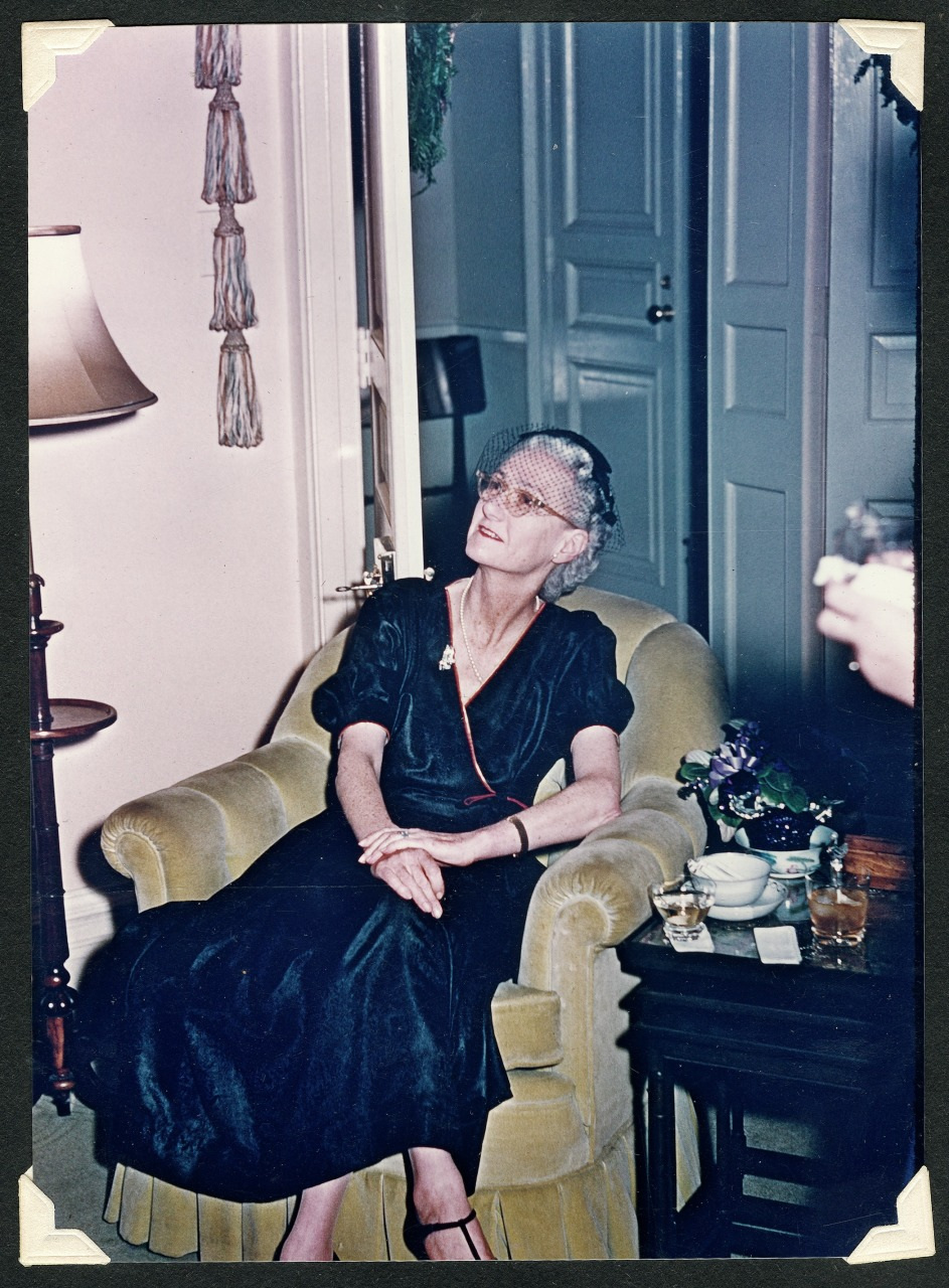 A woman in a dark dress sits elegantly in an armchair, wearing a stylish hat with a veil. She is looking upwards, with her hands resting on her lap. A table with drinks and a potted plant is beside her. The room has a classic interior.