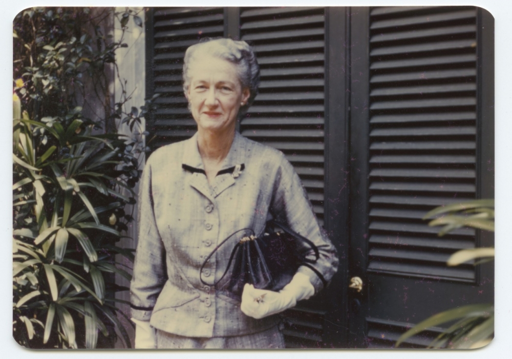 An elderly woman in a gray suit stands outdoors, holding a black purse and wearing white gloves. She is smiling slightly and is positioned in front of dark shutters with green foliage around her.