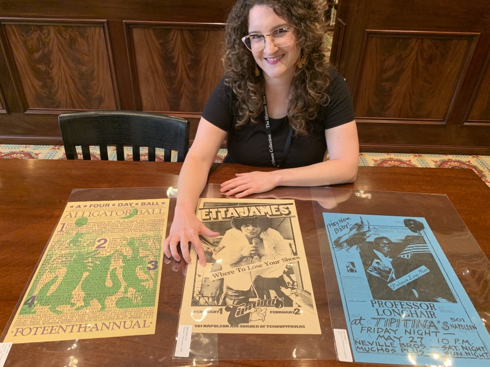 A person sitting at a table smiles while displaying three vintage music posters. The posters feature events like the Alligator Ball, an Etta James concert, and a performance by Professor Longhair. The background includes wooden paneling.