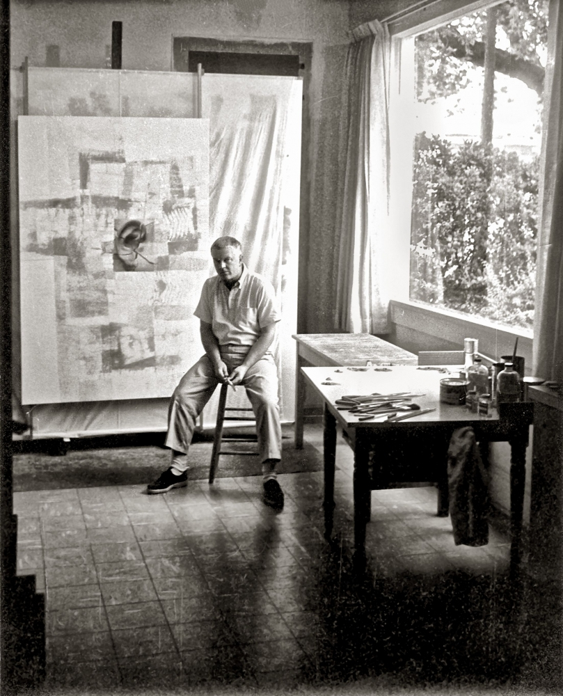 A person sits on a stool in an art studio, surrounded by paintings and art supplies on a table. A large window reveals a view of trees outside. The scene is in black and white.