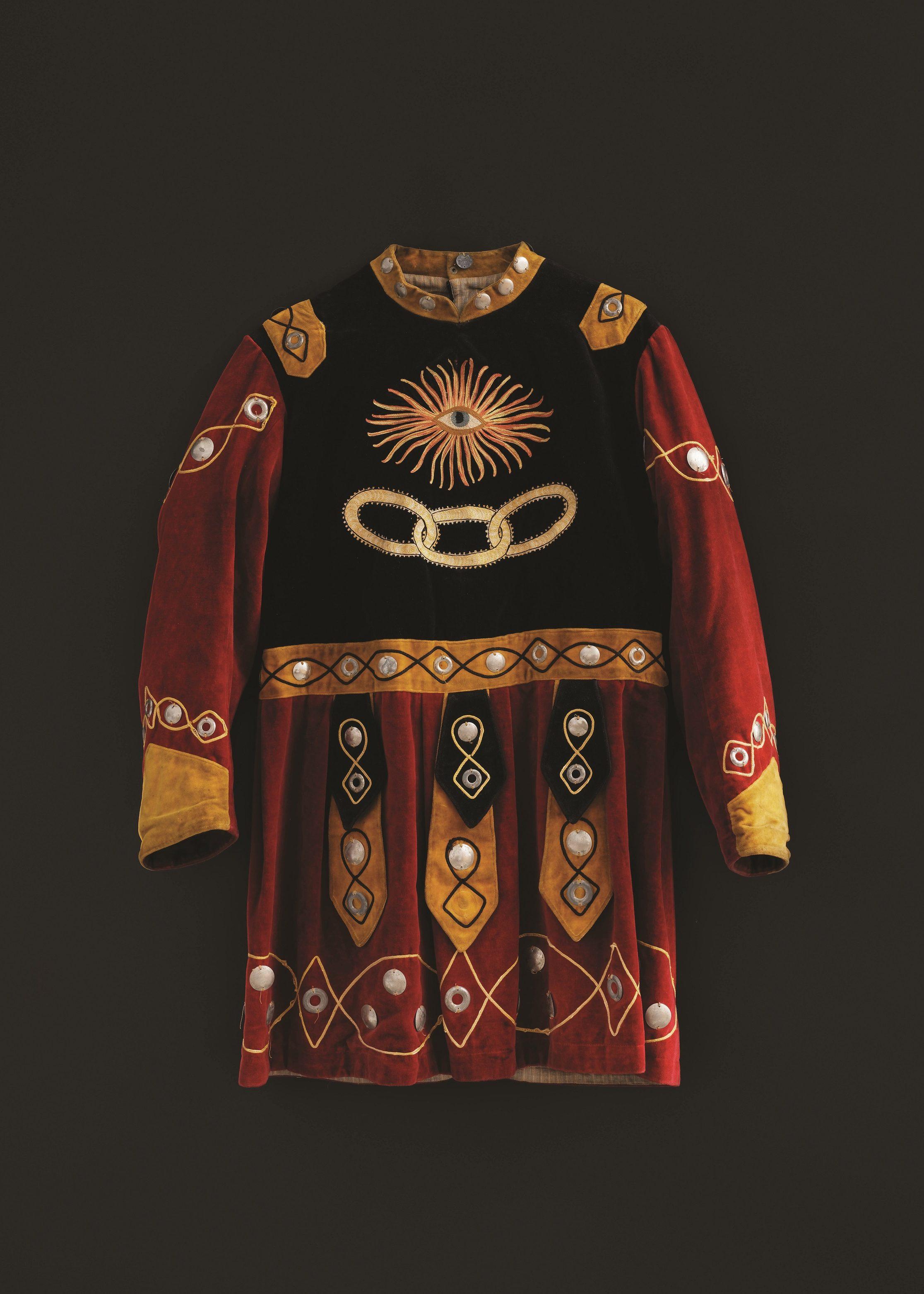 A richly adorned medieval costume with intricate embroidery and patterns in red, black, and gold. The design includes circular and diamond shapes with ornate detailing on the sleeves and bodice, set against a dark background.