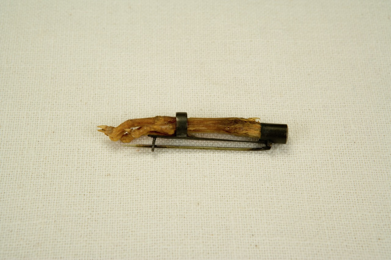 A small, rustic pin resembling a twig is fastened with a metal clamp on a neutral fabric background.