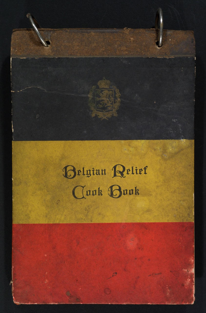 The image shows a vintage Belgian Relief Cook Book with a cover design resembling the Belgian flag, featuring horizontal black, yellow, and red stripes. A decorative emblem is at the top, and the book is bound with metal rings.