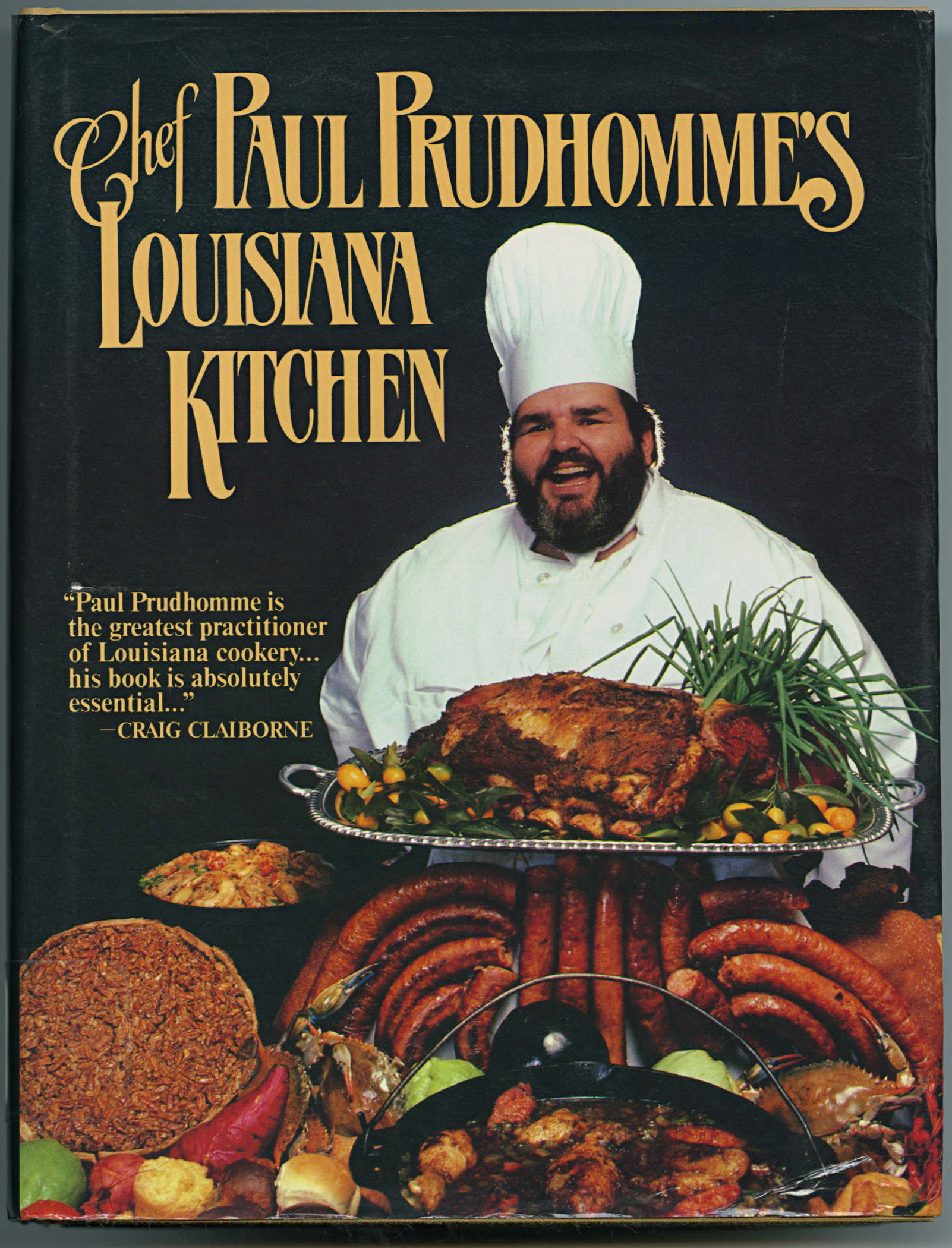 Cover of Chef Paul Prudhommes Louisiana Kitchen features a smiling chef in a white hat and coat. In front of him, theres a display of various cooked meats and vegetables. The background is dark with the title in large, elegant letters.