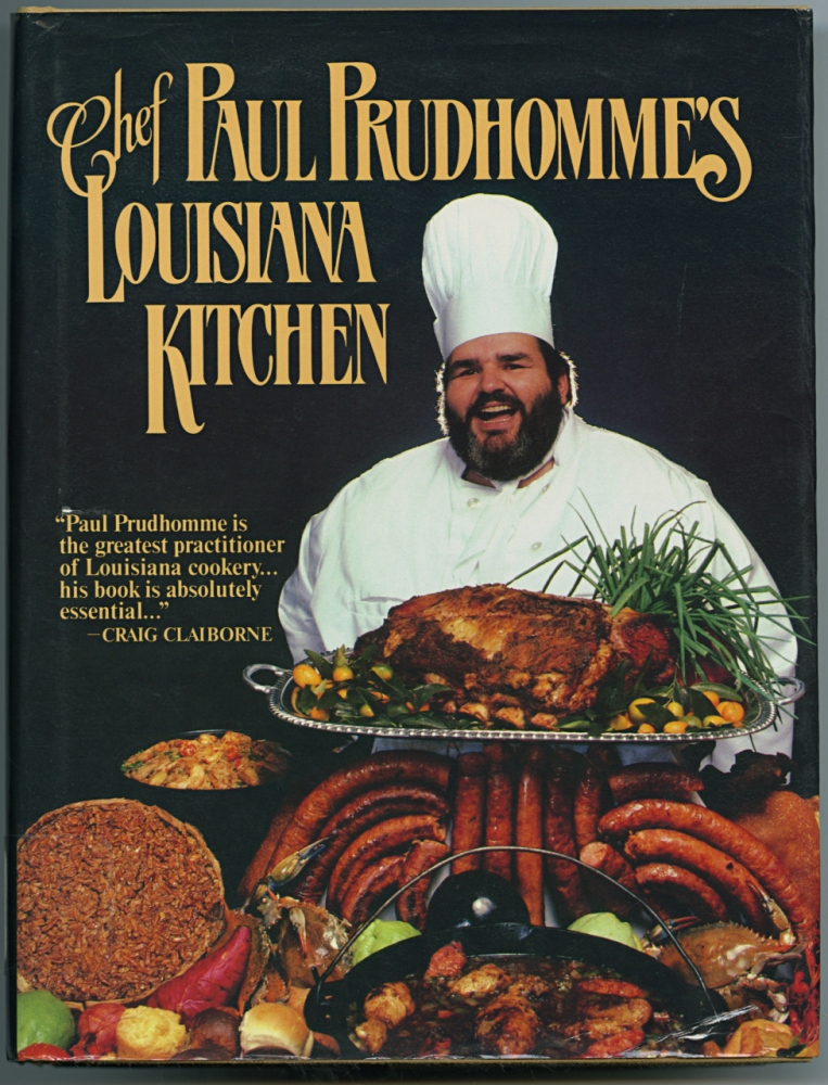 Cover of Chef Paul Prudhommes Louisiana Kitchen cookbook. Features Chef Paul in a white chefs coat and hat, smiling behind a table laden with various Cajun dishes, including meats and vegetables. Text with a quote from Craig Claiborne is also visible.