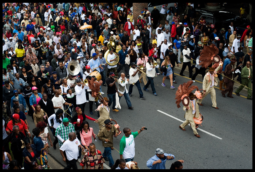 A lively street parade with a large crowd, featuring a brass band playing instruments and two men in feathered costumes. The diverse crowd is walking and enjoying the festive atmosphere.