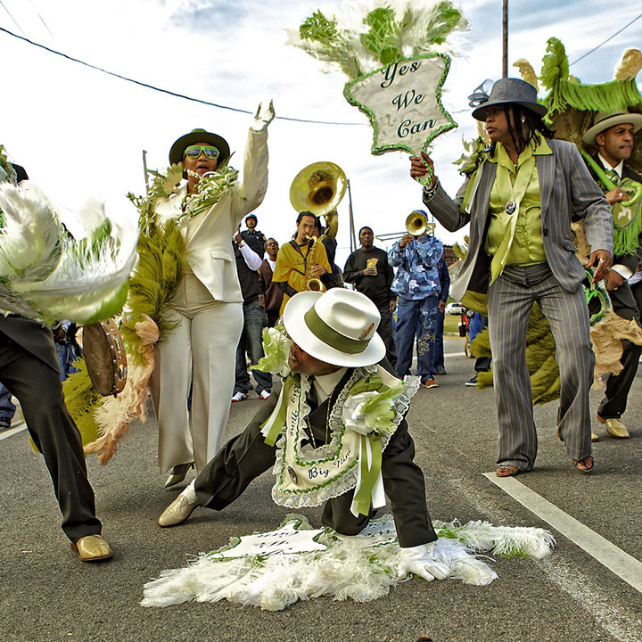 A vibrant parade features individuals dressed in green and white costumes. One person performs a move on the street, while others dance and play instruments. A sign reads Yes We Can in the background. The atmosphere is lively and festive.