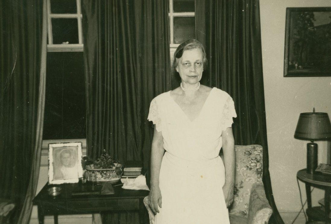 A woman in a white dress stands in a vintage room with dark curtains. A table with a framed photo, a decorative bowl, and books is beside her. A patterned armchair and a lamp with a shade are visible in the dimly lit setting.