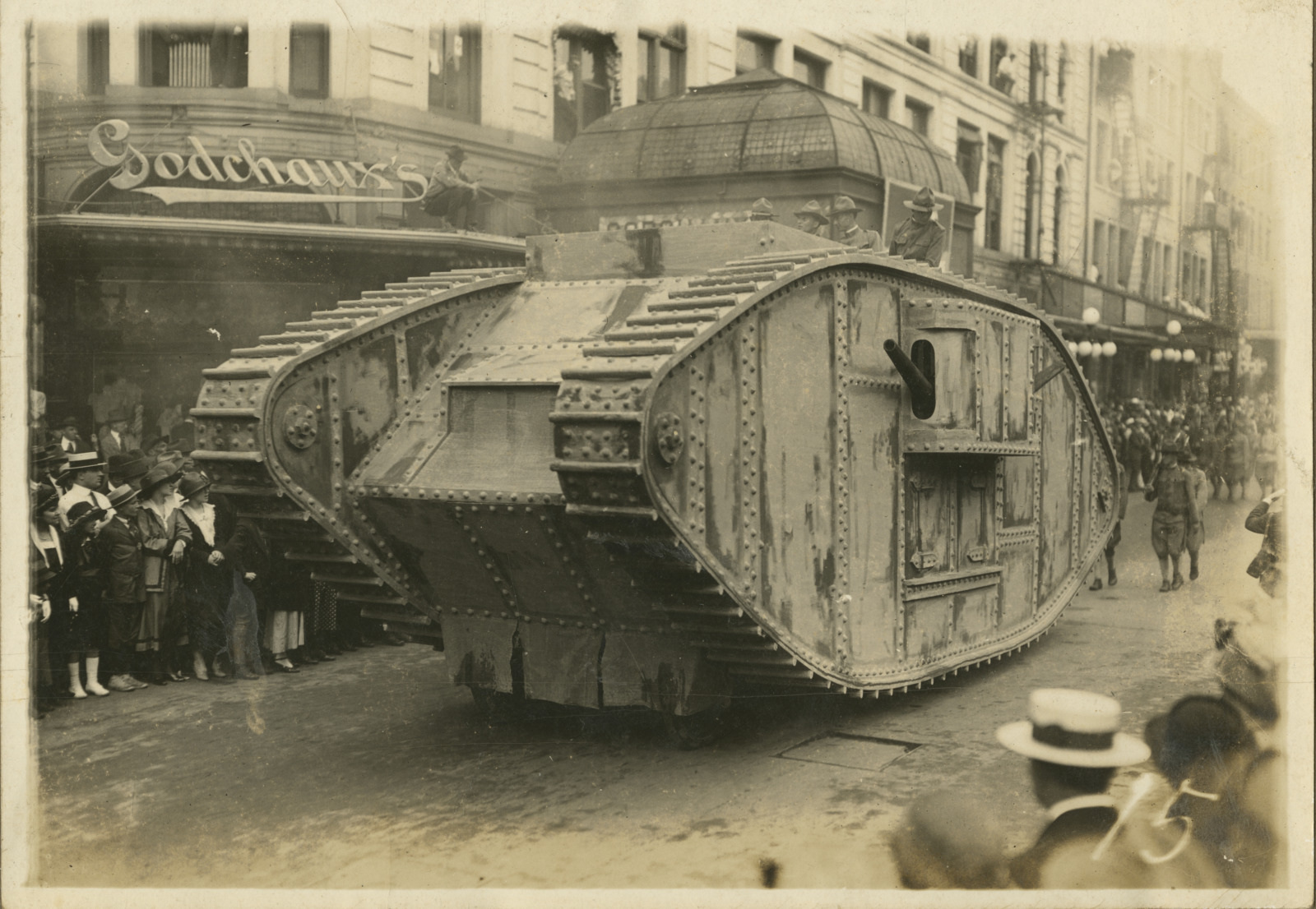 A World War I tank parades down a city street lined with onlookers. The tank, featuring riveted armor, moves past a building with a Godchauxs sign. Spectators, including several in hats, stand on the sidewalk observing the scene.