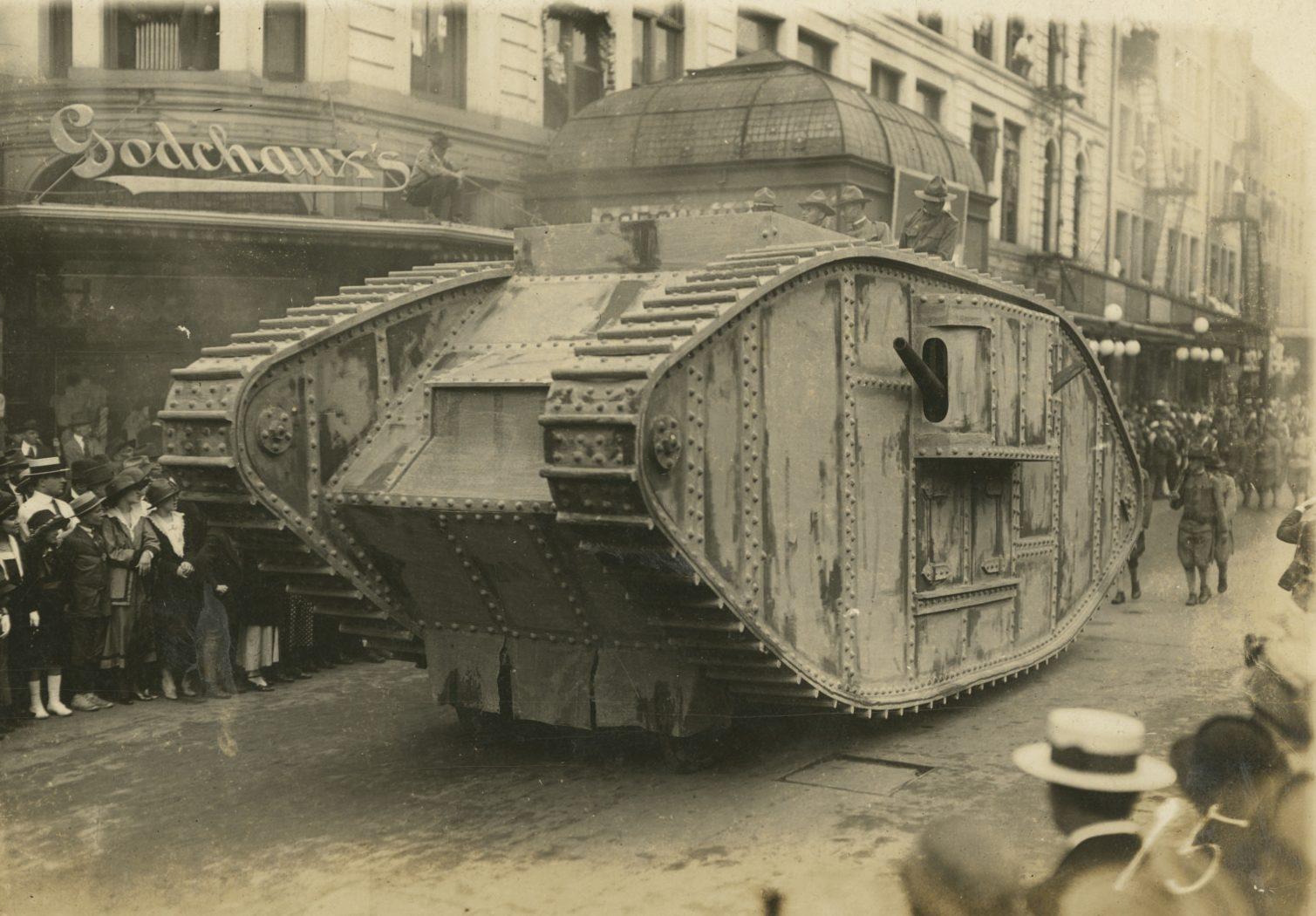 A vintage photograph shows a large military tank on a city street with onlookers, including children and adults in early 20th-century attire. Buildings with signs are visible in the background.