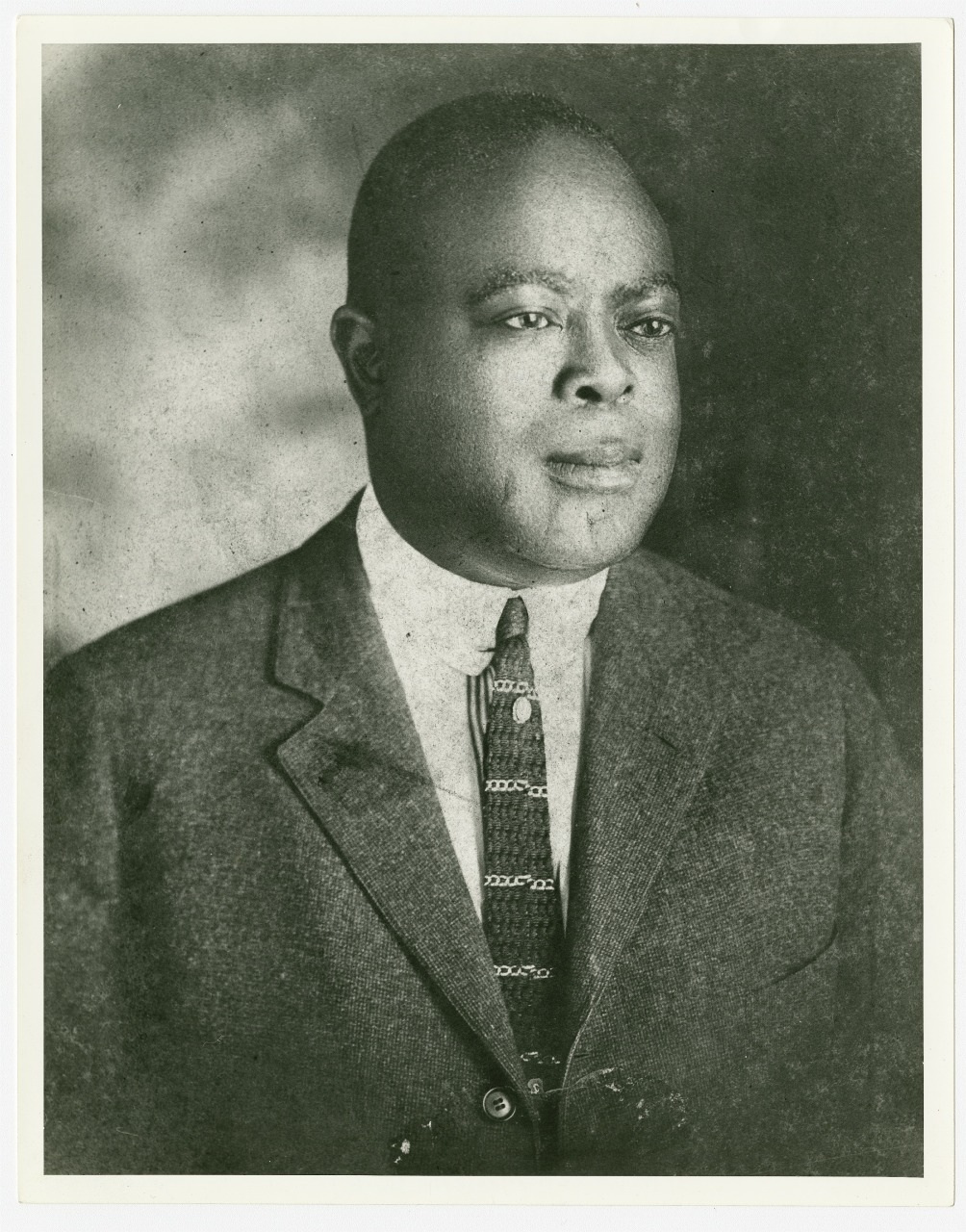 A black and white photo of a man in a suit and tie, gazing slightly to the side. The background is plain, and the image focuses on the mans upper body and facial expression.