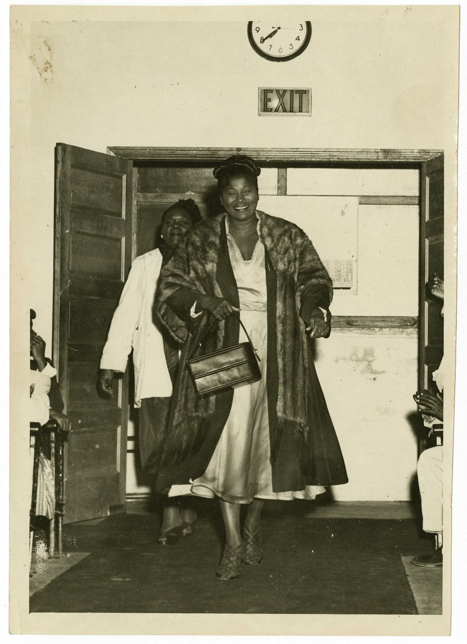 A confident woman in a fur coat and dress walks through a doorway under a wall clock showing 10:09 and an exit sign. Another person follows behind, and two people sit on either side of the entrance.