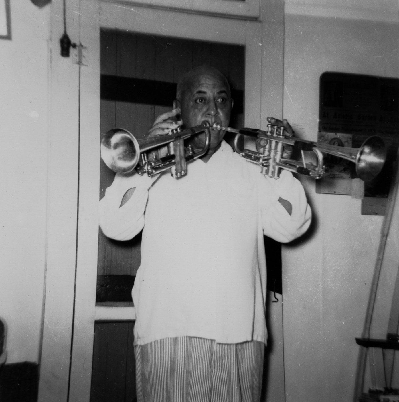 A person playing two trumpets at once indoors, wearing a white shirt and striped pants, standing in front of a door. The room has various items visible in the background, including a sign and other objects. The image is in black and white.