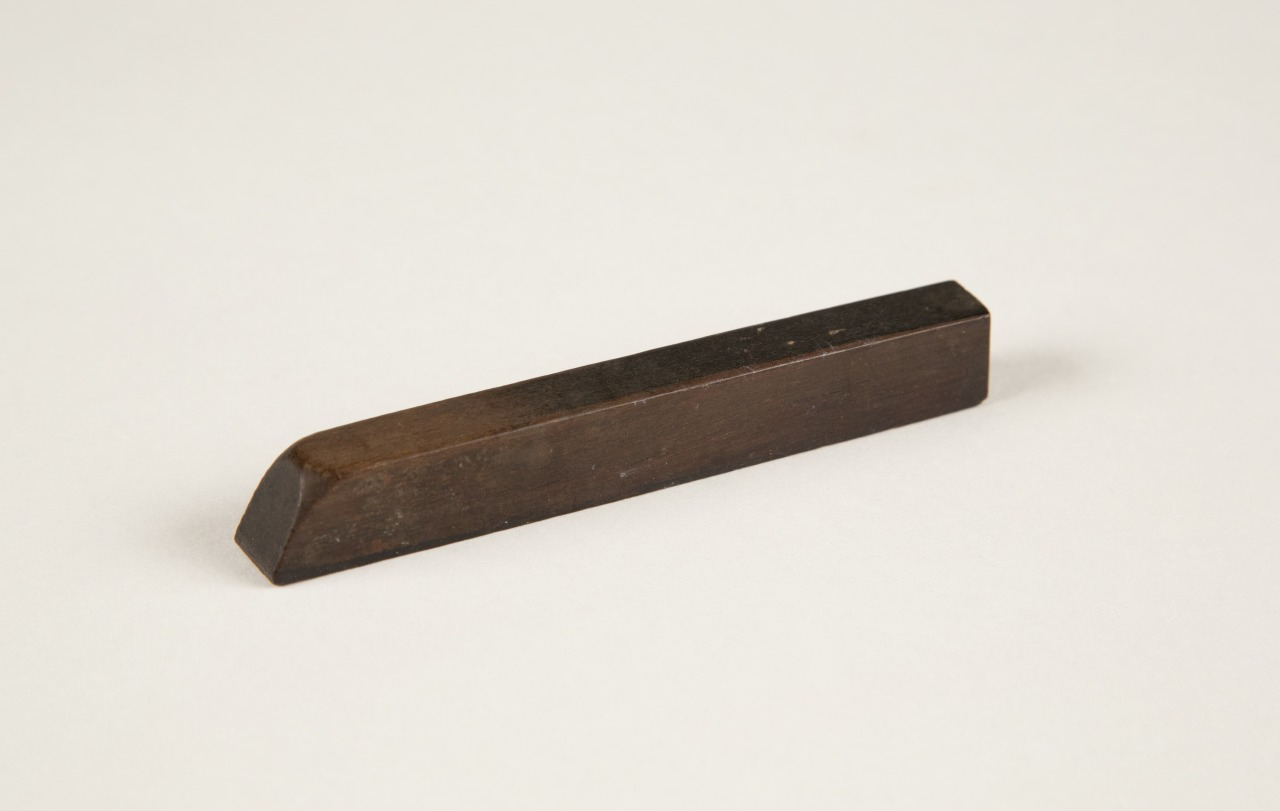 A rectangular metal bar lying on a white surface, featuring a slanted end on one side and a flat end on the other. The metal appears dark and matte, suggesting it might be made of steel or a similar material.