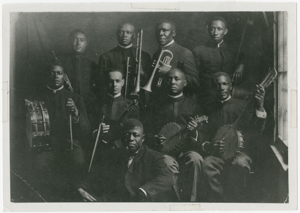 A vintage black and white photo of nine men in formal attire holding musical instruments, including a trumpet, clarinet, drums, banjos, and possibly stringed instruments. They are posed in a group, looking at the camera.