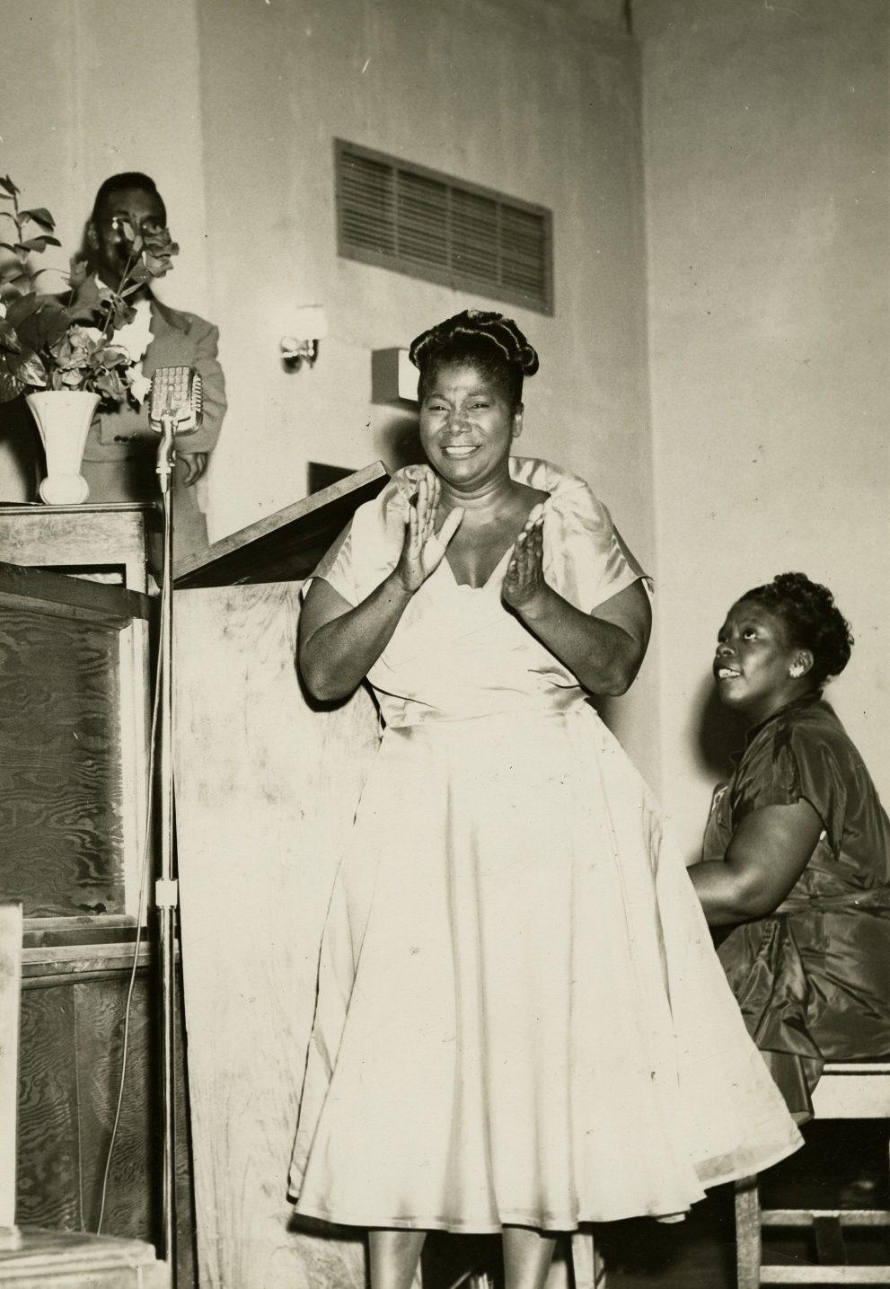 A woman in a light-colored dress stands clapping on stage, while another woman in a dark dress sits at a piano. A man speaks into a microphone behind a podium with flowers on it. The setting appears to be a church or hall.