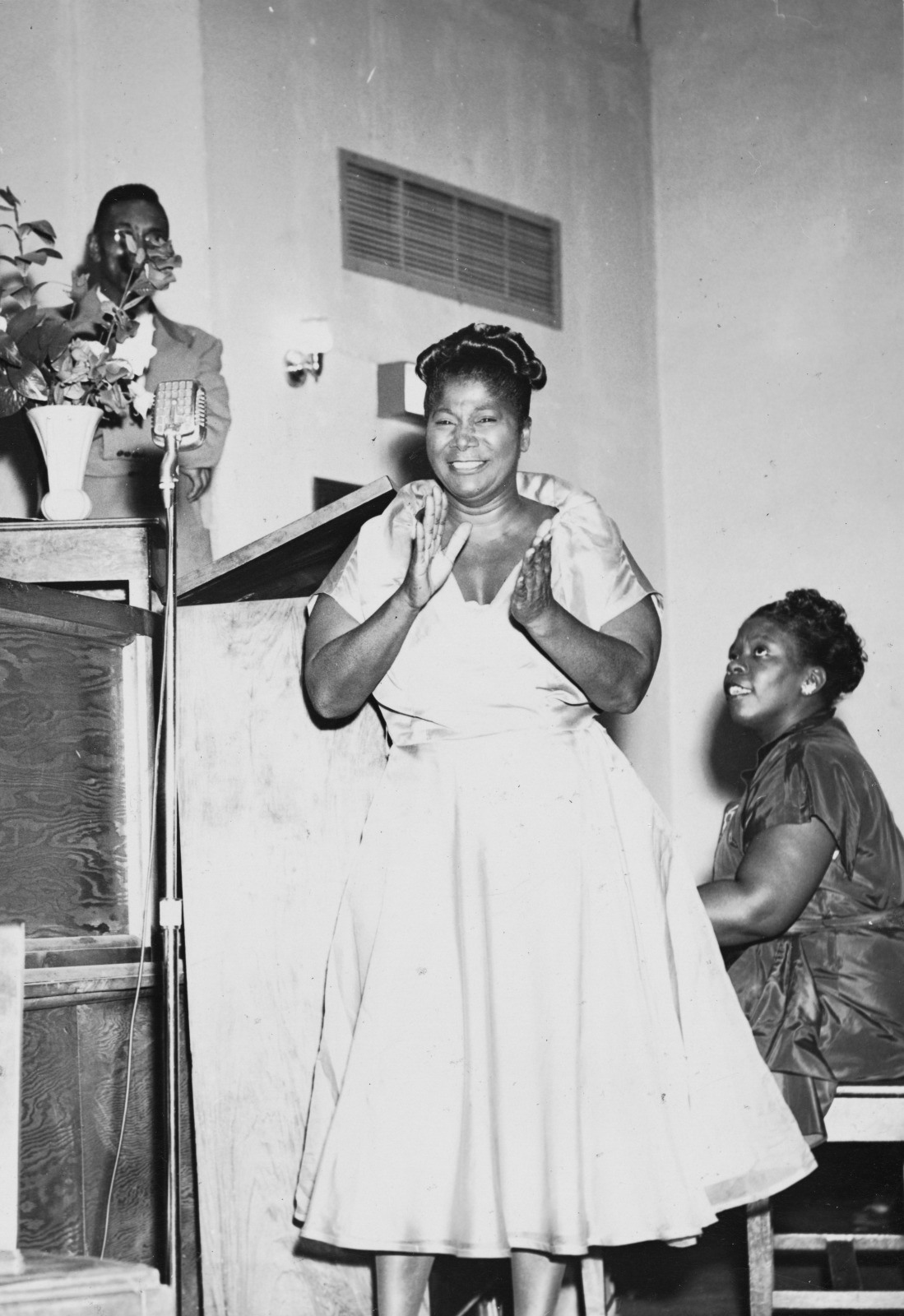 A woman in a dress claps her hands while another woman plays the piano beside her. A man stands at a podium in the background. They appear to be in a church or auditorium setting.