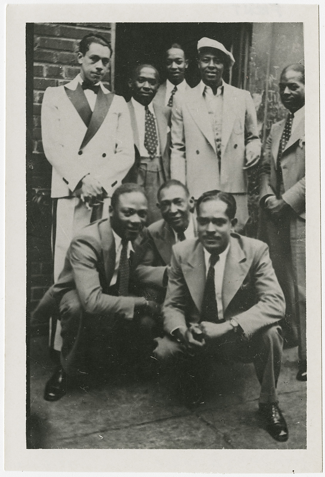 A vintage black and white photo of seven men in suits. Five are standing, with one wearing a white suit and fedora. Two are crouching in front. They are posing outdoors against a brick wall and a doorway. The style suggests an early to mid-20th century setting.
