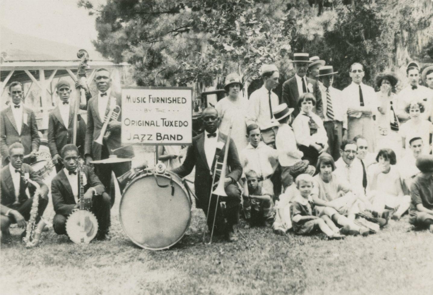 A vintage black-and-white photo of the Original Tuxedo Jazz Band. The group, including men and women, is gathered outdoors with musical instruments. A sign reads Music Furnished by the Original Tuxedo Jazz Band.