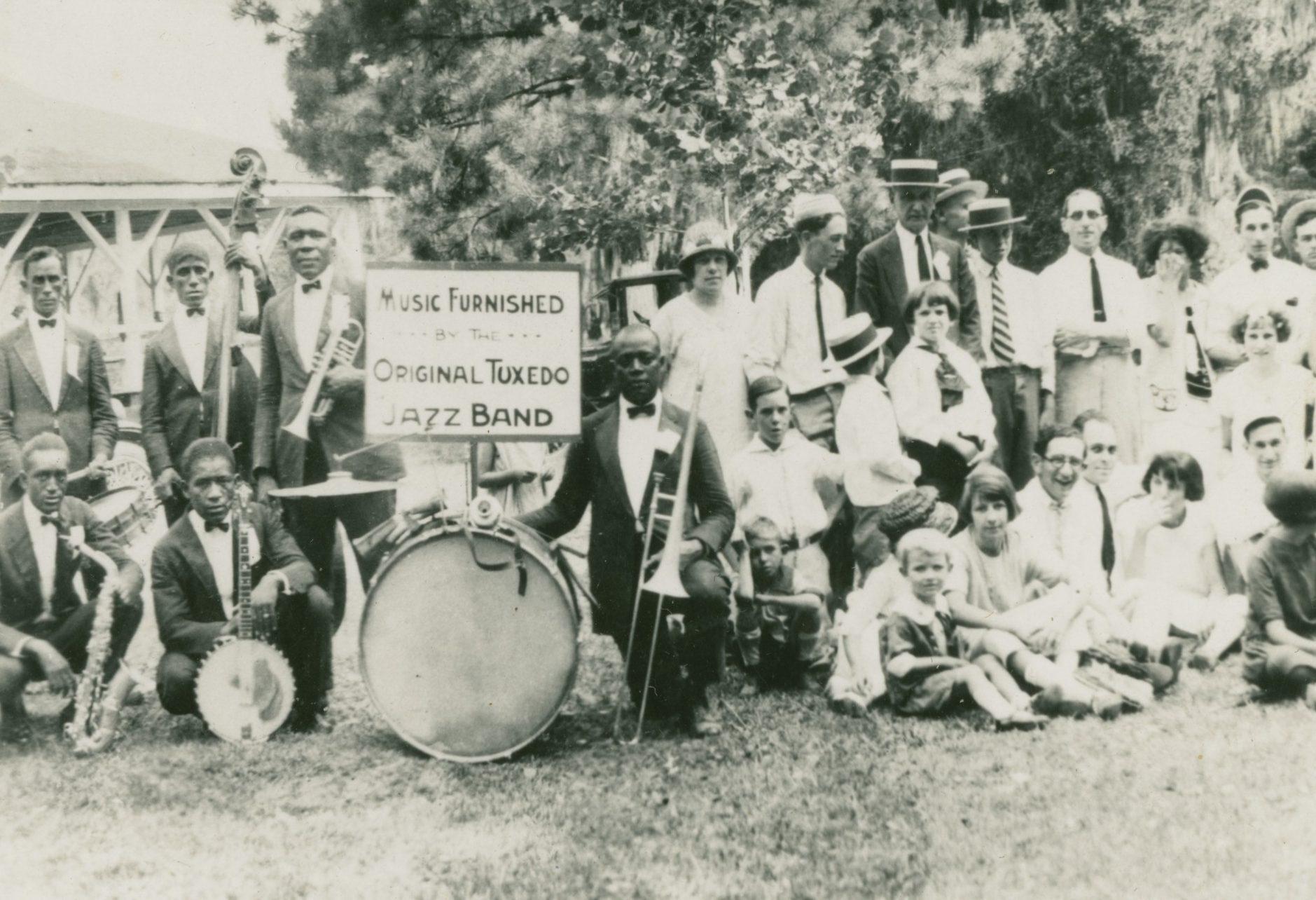 A vintage black and white photo of a jazz band with various members, including musicians holding instruments like trumpets and drums. A sign reads Music Furnished by the Original Tuxedo Jazz Band. A group of people gather around, some seated on grass.