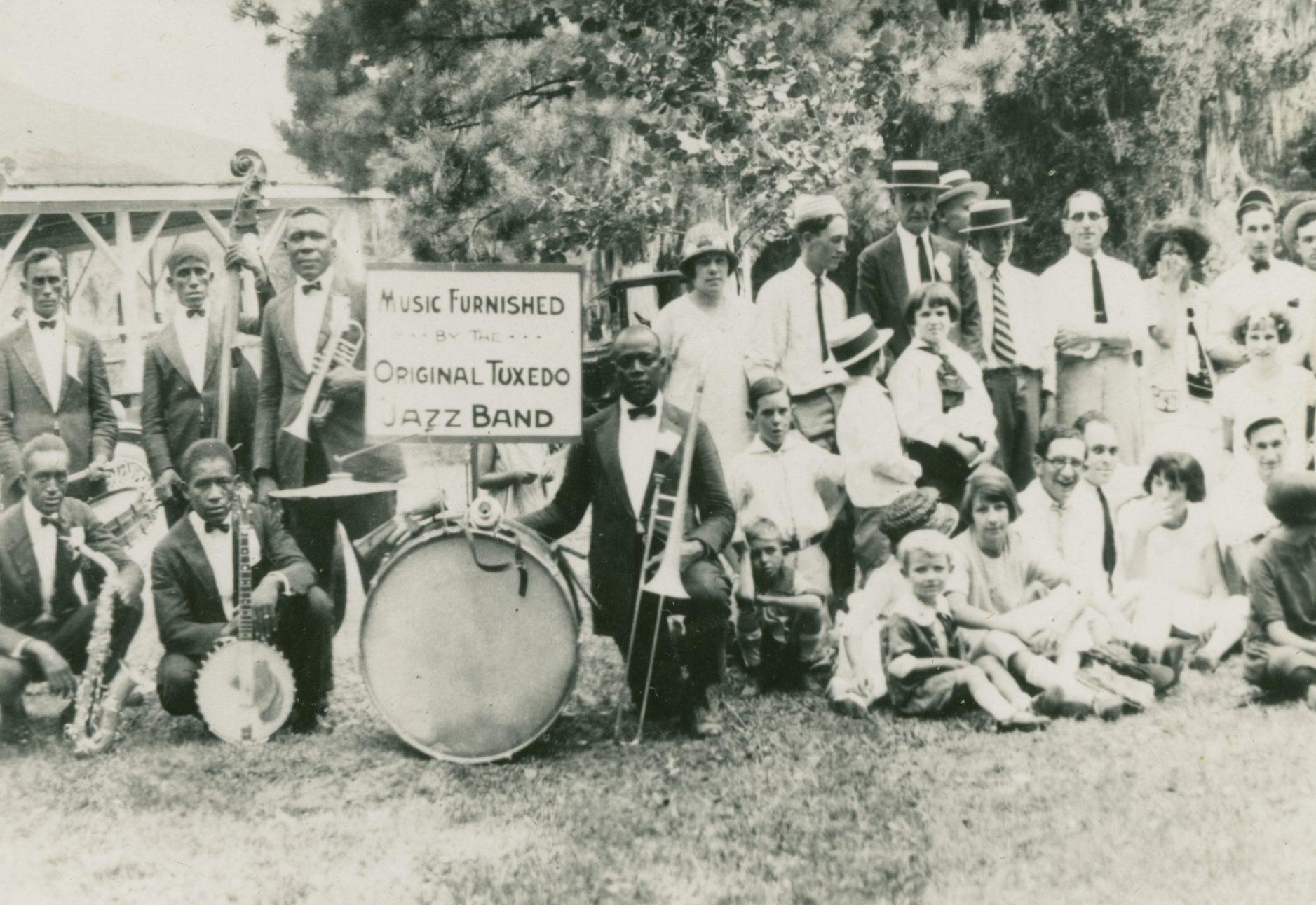 A black and white photo of the Original Tuxedo Jazz Band posing outdoors with their instruments. A banner reads Music Furnished by the Original Tuxedo Jazz Band. Many people, some seated and some standing, are gathered around them.
