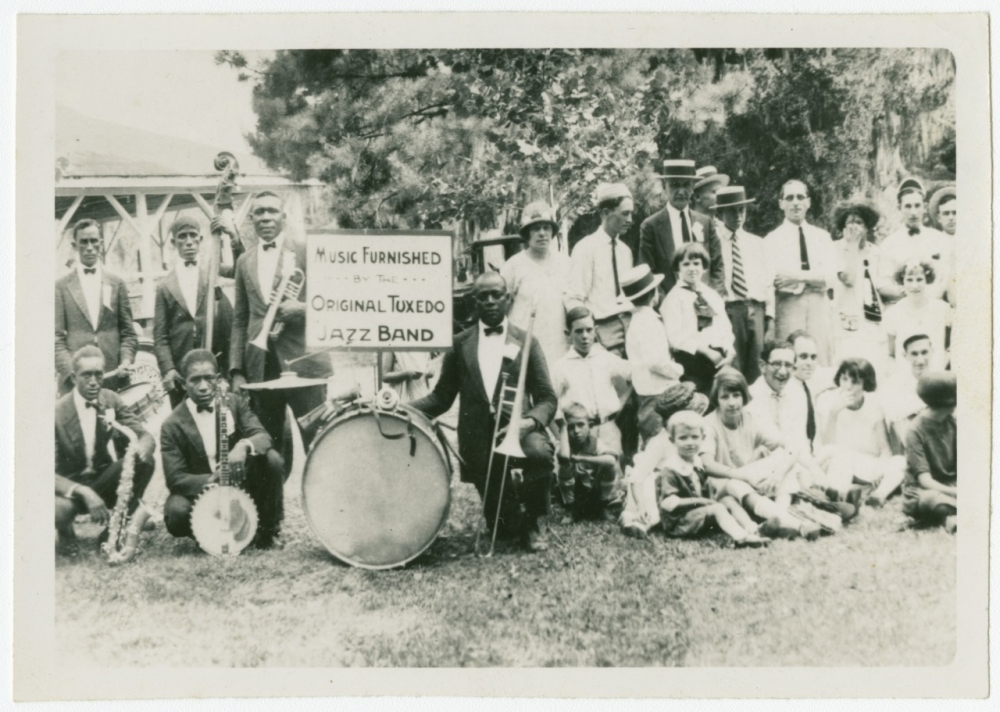 A black and white photo of a jazz band and a group of people outdoors. Musicians hold instruments, including a saxophone and a drum with a sign reading Music Furnished By The Original Tuxedo Jazz Band. The setting appears casual and lively.