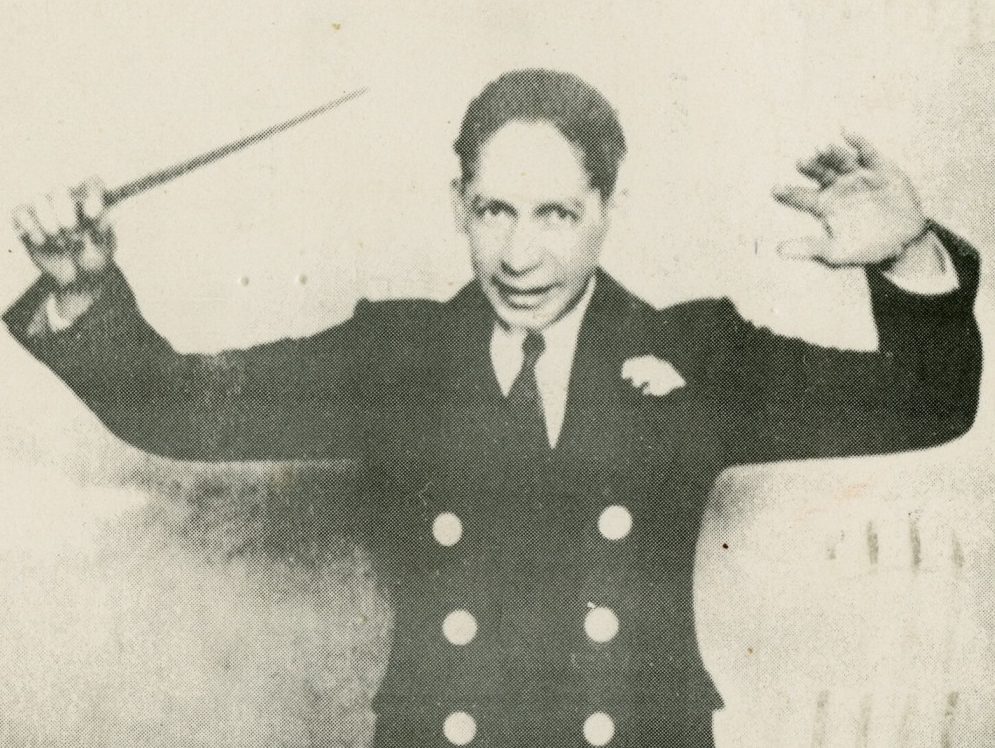 A man in a double-breasted suit gestures with both hands, holding a baton in his right hand. The black-and-white image gives a vintage feel, with the man expressing a dynamic and commanding presence.