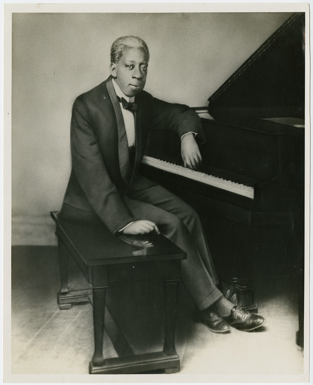 A vintage black and white photo of a person seated at a piano, wearing a dark suit and bow tie. They have a composed expression and are leaning casually against the piano with one arm resting on the keys.