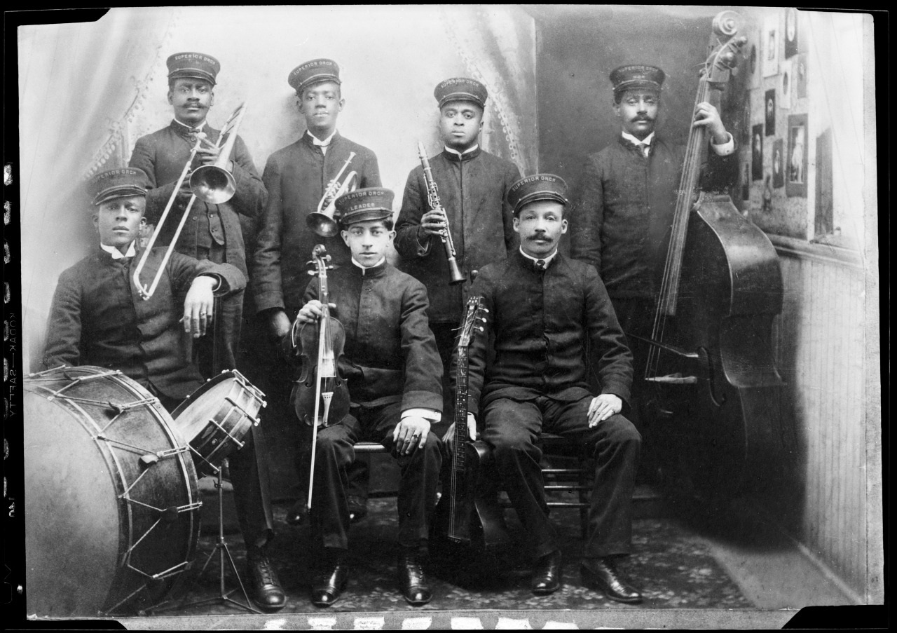 A vintage black and white photo of a seven-member band. The musicians are dressed in uniforms and hold various instruments including a drum, trombones, violins, clarinet, and double bass. They are seated and standing in a formal pose against a plain backdrop.