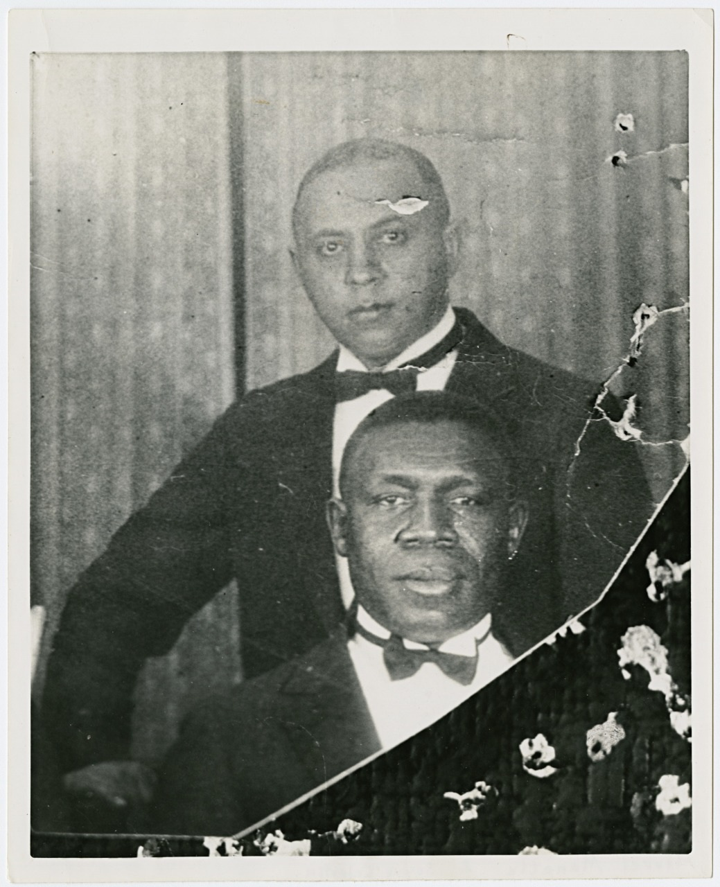 Two men in formal attire pose for a vintage black and white photograph. The man in front is seated, and the man slightly behind is standing. Both wear tuxedos with bow ties. The photo shows signs of aging with visible scratches and creases.