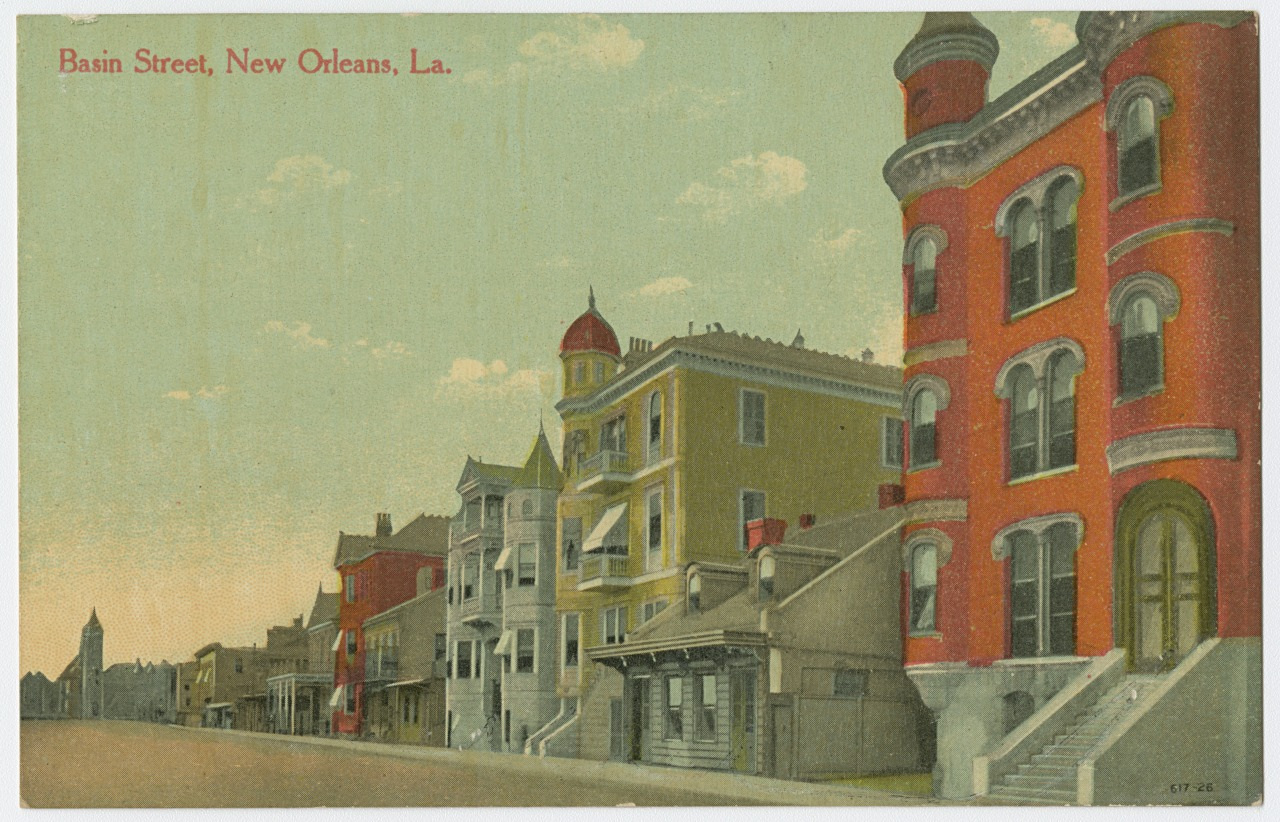 A vintage postcard depicting a street view of Basin Street in New Orleans, Louisiana. The image shows a row of colorful, ornate buildings, including a prominent red brick building with round towers. The sky is lightly clouded.