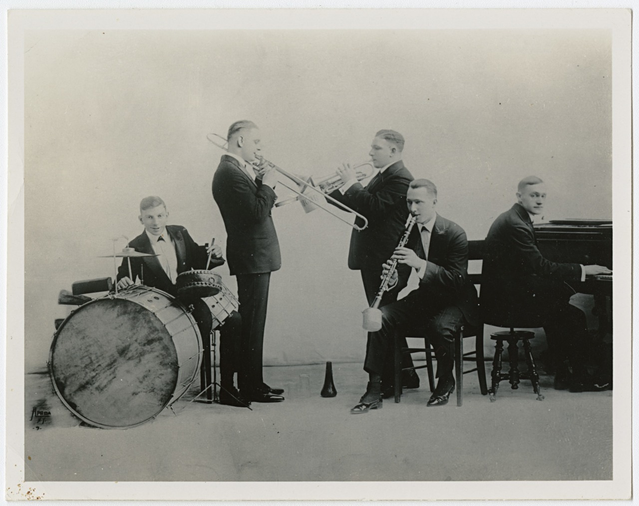 A vintage black-and-white photo of five musicians in suits. One plays the drum, another a trombone, one a trumpet, another a clarinet, and one plays the piano. They are arranged in a semi-circle, focused on their respective instruments.