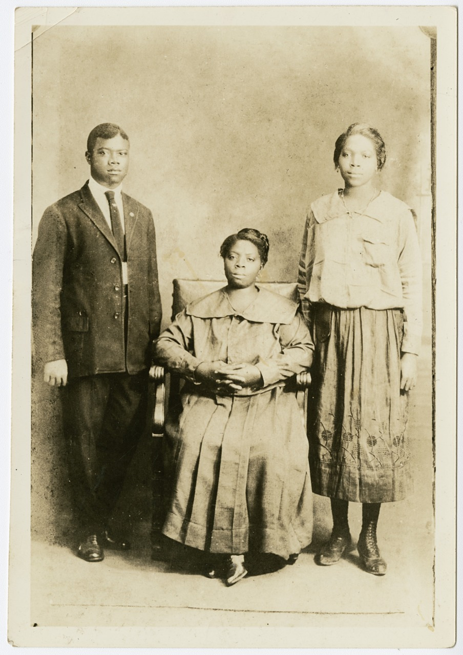 A vintage black and white photograph shows three people. A seated woman in a long dress is in the center, with a young man in a suit on her left and a young woman in a blouse and skirt on her right. The background is plain.