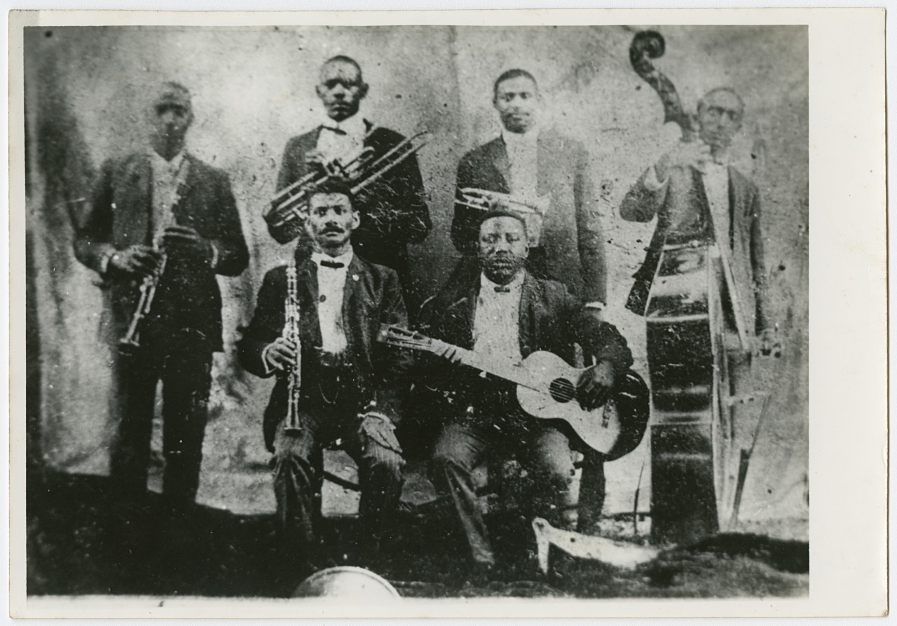 A vintage black and white photo of a six-member jazz band. The musicians are posing with their instruments: a clarinet, trombone, trumpet, guitar, and double bass. They are dressed in suits and standing against a plain backdrop.