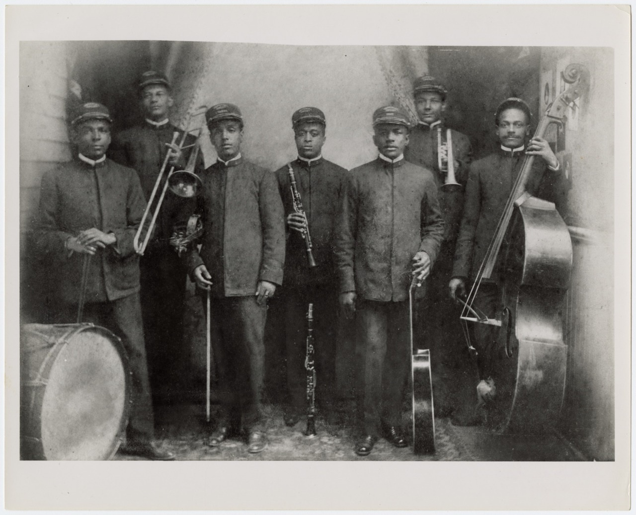 A vintage black-and-white photograph of seven men in uniforms holding musical instruments. The group includes a trombone, trumpet, clarinet, violin, accordion, and double bass. A drum is visible on the left. They pose in front of a curtain.
