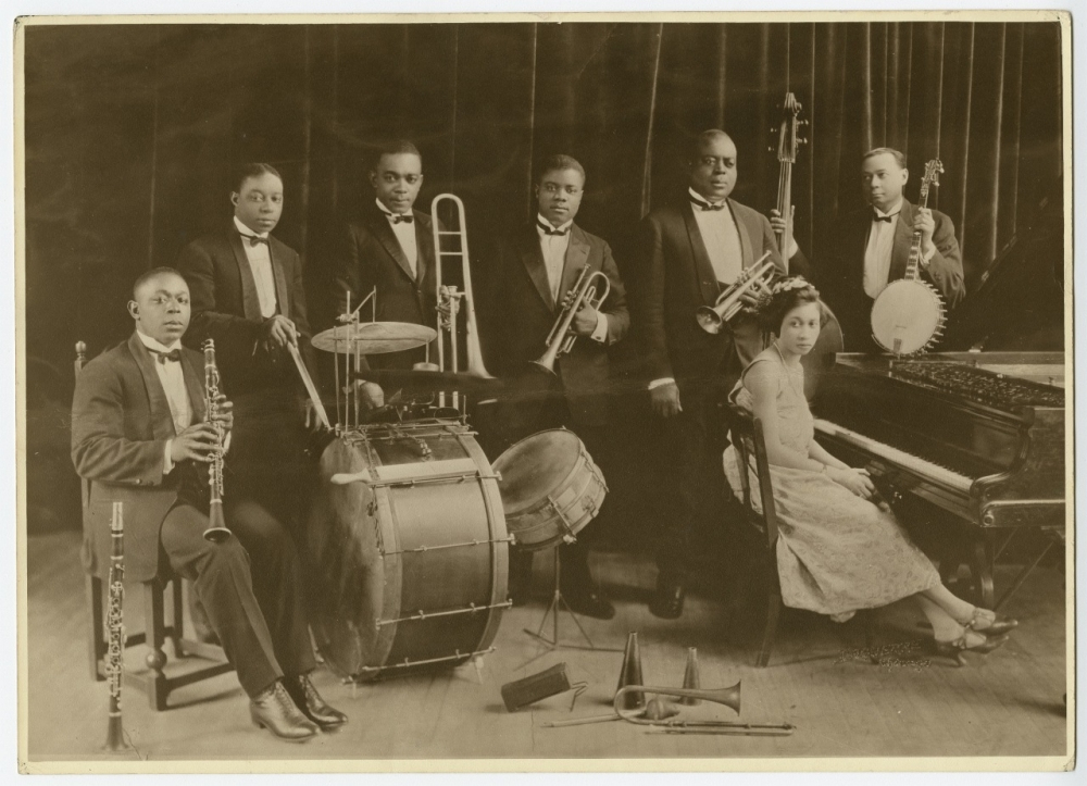 A vintage black and white photograph of a jazz band with seven musicians. Six men stand with instruments like trombone, trumpet, clarinet, banjo, and drums. A woman sits at the piano. They are dressed in formal attire against a dark curtain backdrop.