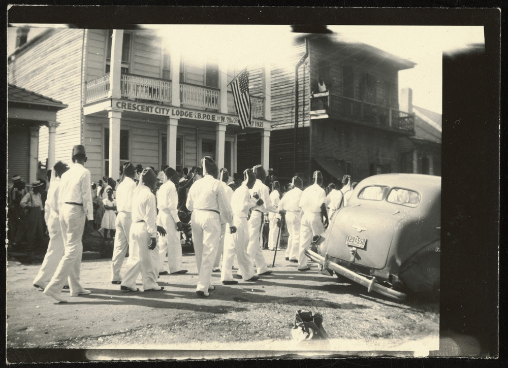 A black and white photo of a group of people in white uniforms marching down a street. They are passing by the Crescent City Lodge building, with an American flag displayed. A vintage car is parked nearby, and onlookers watch from the sidewalk.