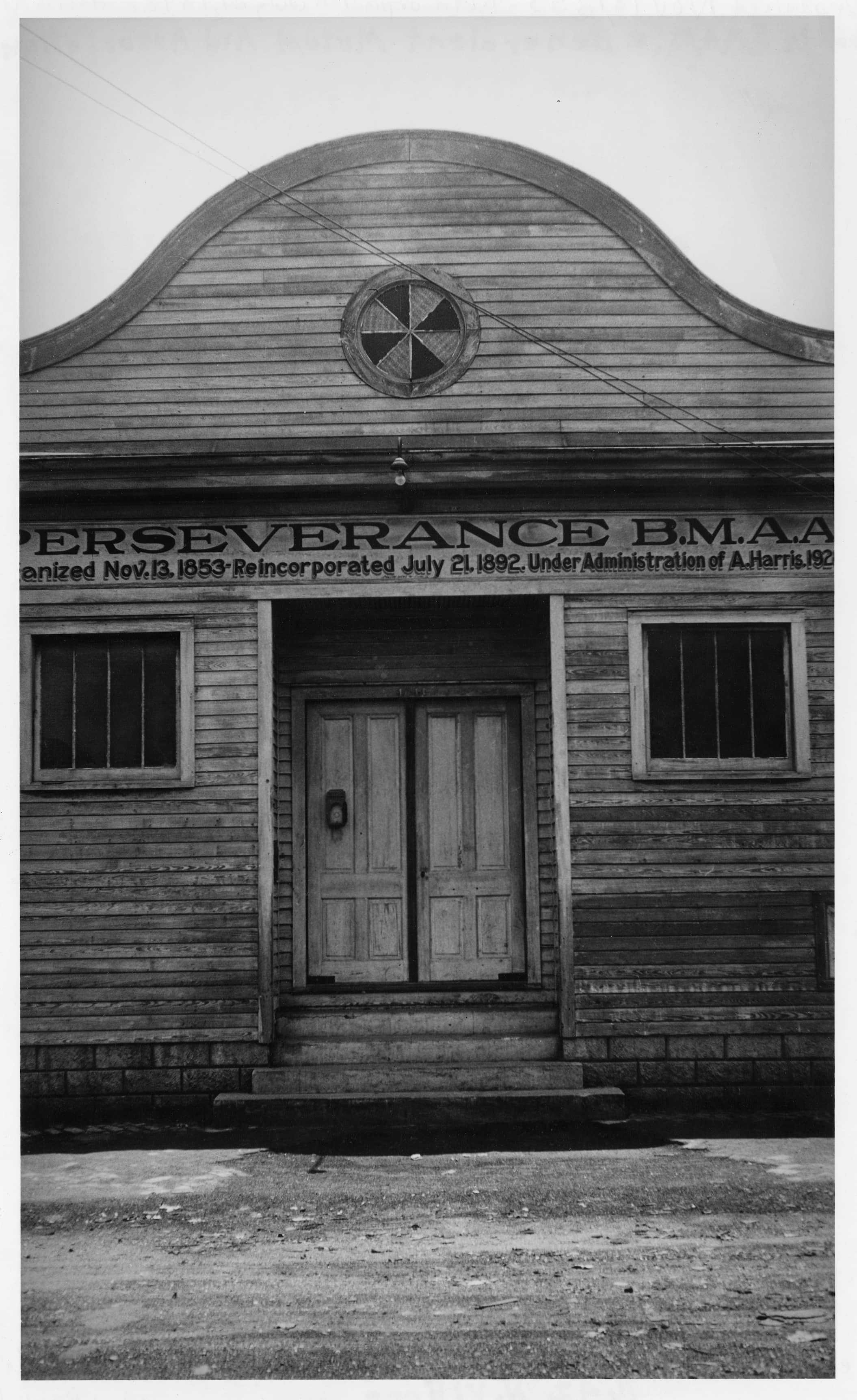 A black and white photo of an old wooden building with a curved facade, two small windows, and a central door. Above the door, a sign reads Perseverance B.M.A.A. with historical dates and text. The building appears weathered.